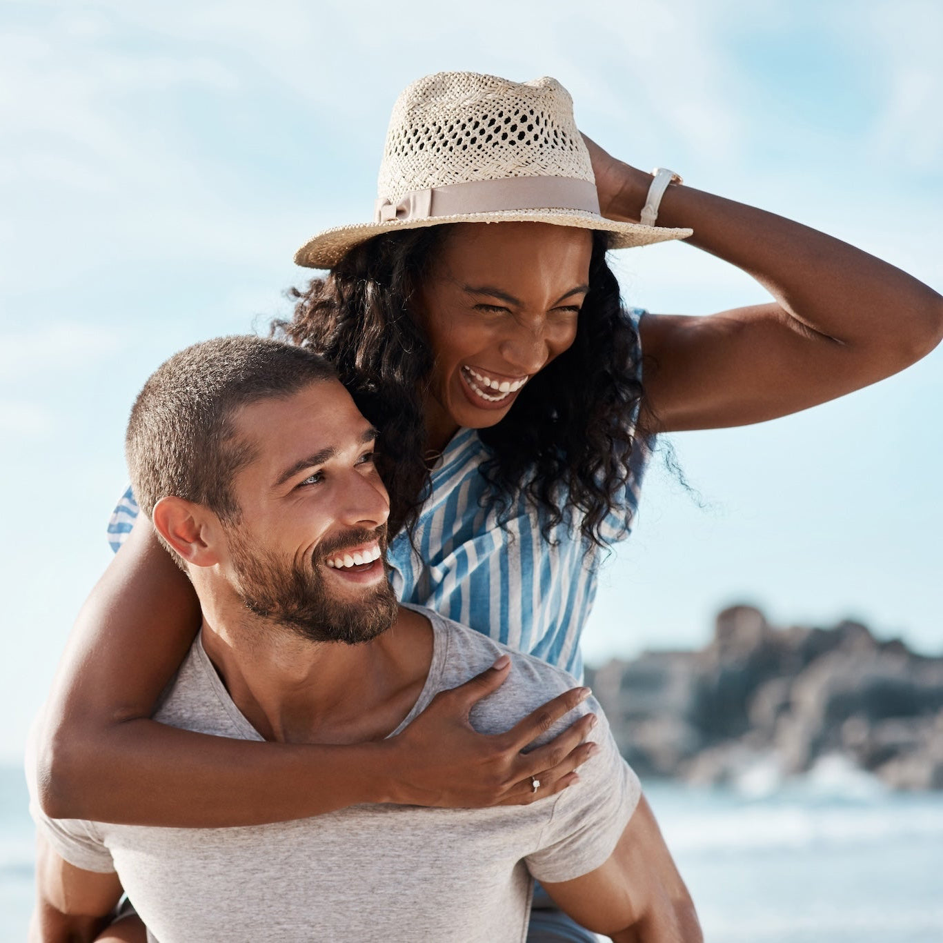 Man giving a piggyback ride to a woman on a beach with a clear sky.